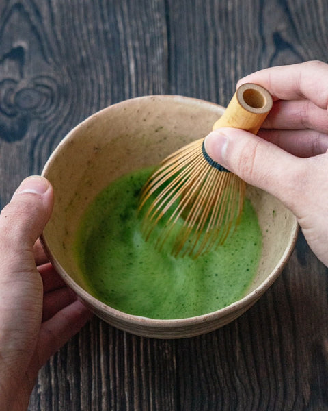 A hand whisking bright-green matcha with a bamboo chasen in a tan ceramic bowl.