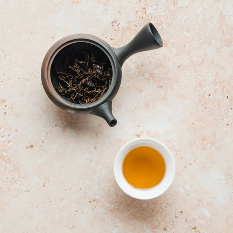 Overhead view of a dark ceramic teapot holding dry Japanese black tea leaves next to a white cup of brewed amber tea on a pale stone surface.