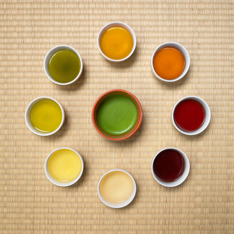 Overhead circular arrangement of small bowls of brewed Japanese teas ranging from pale green to deep amber and red, with a green tea in the center.