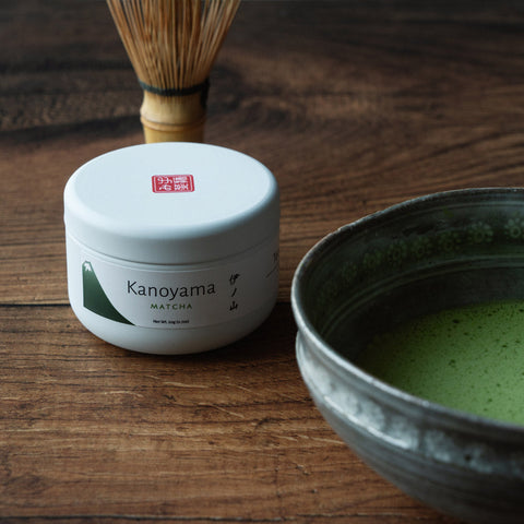 A labeled matcha canister beside a dark bowl of bright-green matcha powder on dark wood.