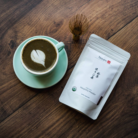A houjicha latte with latte art in a mint-green cup and saucer beside a foil tea pouch and a bamboo whisk on dark wood.