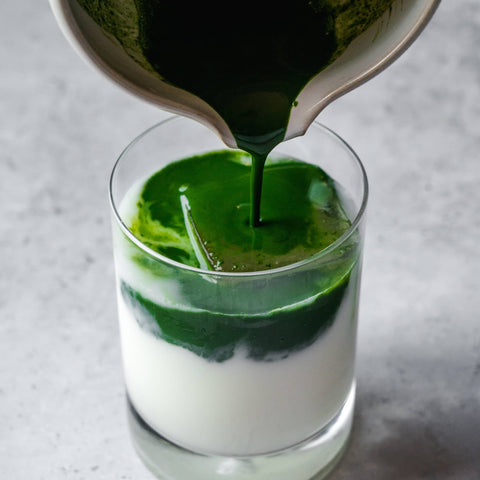 Bright-green matcha being poured over milk into a glass to make a latte on a grey surface.