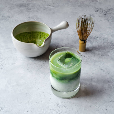 A layered iced matcha latte in a glass with milk below and bright-green matcha on top, with a bowl of whisked matcha and a bamboo whisk behind.