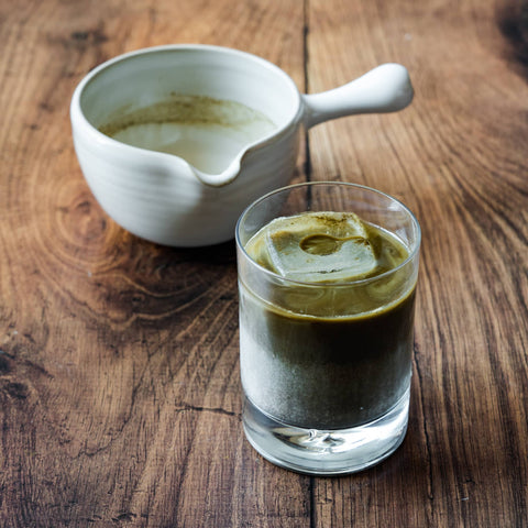 A white lipped pouring bowl beside a glass of layered iced houjicha latte of milk and brown roasted tea on wood.
