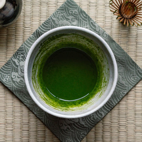 Overhead view of thick, glossy bright-green koicha matcha in a grey stone bowl resting on a tatami mat.