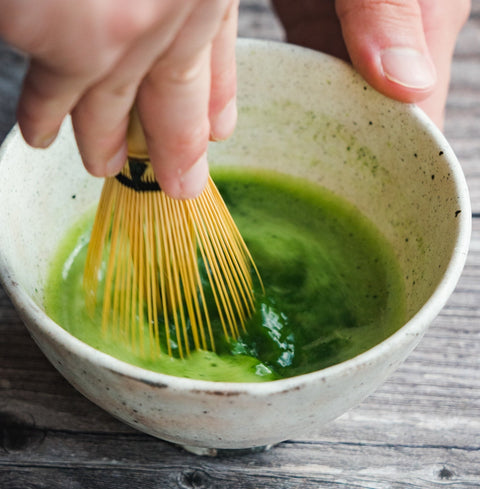 A hand whisking bright-green matcha with a bamboo chasen in a cream bowl, viewed from above.