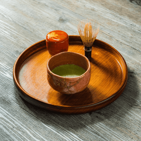 A brown ceramic cup of matcha with a bamboo whisk and a small orange dish on a round wooden tray.