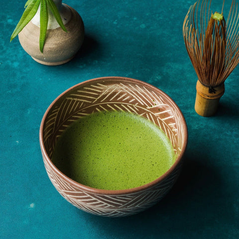 A carved, patterned chawan of bright-green matcha on a teal surface with a bamboo whisk and whisk holder behind.