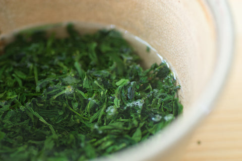 Close overhead view of wet, steeped bright-green sencha leaves in a pale ceramic vessel.