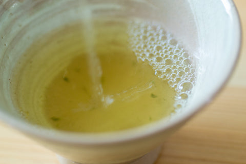 Close overhead view of a pale ceramic cup filled with pale yellow-green brewed tea showing fine foam on the surface.