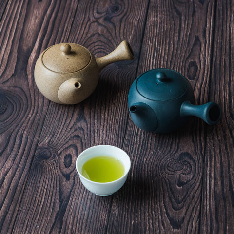A tan and a dark teal Japanese kyusu teapot beside a cup of brewed green tea on a dark wooden surface.