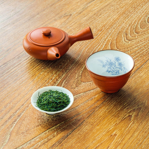 A red-clay Tokoname kyusu side-handle teapot beside a cup of brewed green sencha and a small dish of dry sencha leaves on a wooden table.