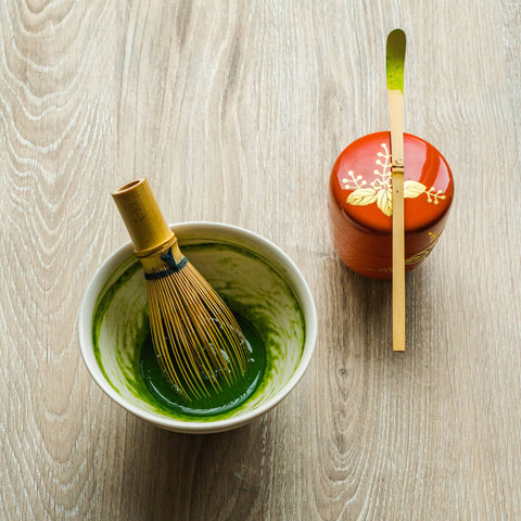 Overhead view of a dark bowl of whisked bright-green matcha with a bamboo chasen whisk, beside a red dish holding a bamboo scoop of matcha powder.