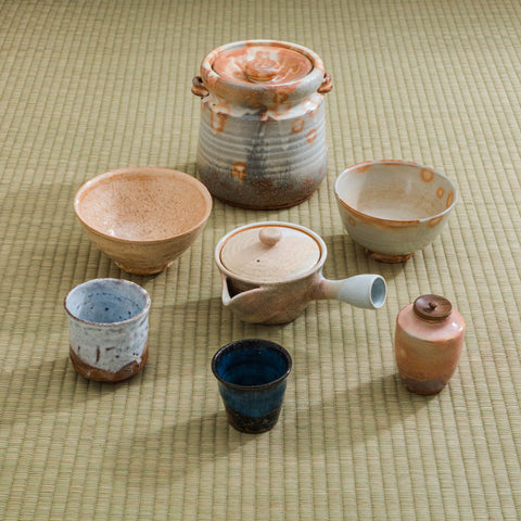 A collection of pale Hagi-yaki teaware including a lidded jar, cups and small bowls in cream, pink and blue glazes arranged on a straw mat.