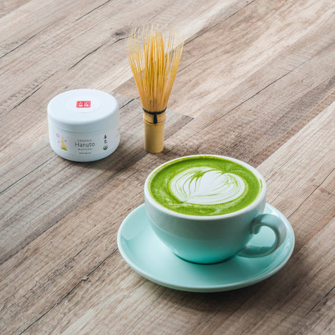 A matcha latte with leaf latte art in a mint-green cup and saucer, with a small tin and a bamboo whisk on a wooden table.