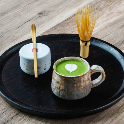 Overhead view of a ceramic mug of matcha latte with heart latte art on a black tray, with a bamboo whisk and small dish.