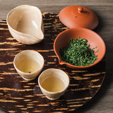 Overhead view of a gyokuro brewing set on a dark woven mat with cream cups, a red-clay bowl of fresh green leaves and a pouring vessel.