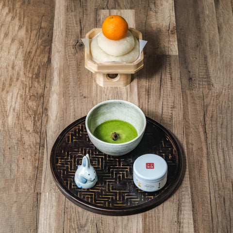 A black tray set with a matcha bowl, small dishes and a New Year ornament topped with an orange fruit.