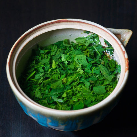 Close-up of an orange-and-blue drip-glazed bowl filled with freshly plucked bright-green tea leaves.