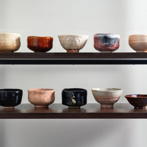 A shelf display of many matcha bowls in assorted glazes and colors, including black, cream and red, arranged on two wooden shelves.