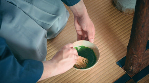 Overhead view of hands whisking matcha in a ceramic bowl while seated on tatami
