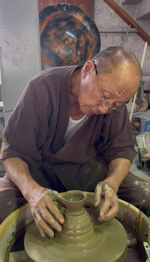 An elderly potter in a brown work kimono throwing a small lidded form on the wheel, a large glazed platter hanging on the workshop wall behind him