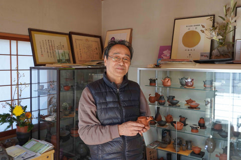 Fugetsu holding a ceramic pot in a room with shelves displaying various ceramic items.