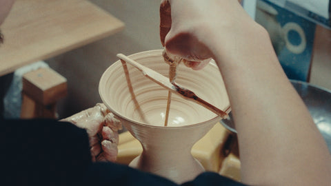 A hand steadying a white porcelain bowl while a brush sweeps glaze across its interior