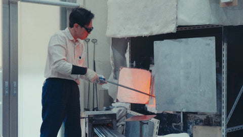Glassblower in dark goggles drawing a gather of molten glass from the glowing furnace