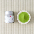 Silver Tezumi tin of Betsugi (別儀) matcha beside a small dish of green matcha powder on a tatami mat.