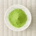 Green Eiryoku matcha powder mounded in a white dish on a tatami mat, overhead view.
