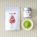 Tezumi Hika matcha pouch with red flame artwork beside a silver tin and a small dish of green matcha powder on a tatami mat.