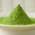 Bright green Hika matcha powder heaped in a tall cone in a cream ceramic bowl, side view.