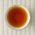 Cup of brewed Honyama Yabukita wakoucha showing a bright amber-orange liquor, shot from above on tatami