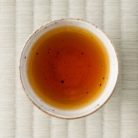 Cup of brewed Honyama Yabukita wakoucha showing a bright amber-orange liquor, shot from above on tatami