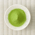 Green Komiya matcha powder mounded in a white dish, viewed from above on a tatami mat.