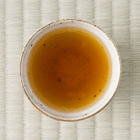 Kumamoto Zairai wakoucha brewed to a deep amber liquor in a speckled white cup, shot from overhead on tatami