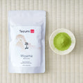 Tezumi Miyama matcha 100g pouch with a misty mountain design beside a small dish of green matcha powder on a tatami mat.