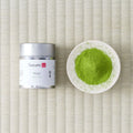 Silver Tezumi tin of Rikyu matcha beside a small dish of green matcha powder on a tatami mat.