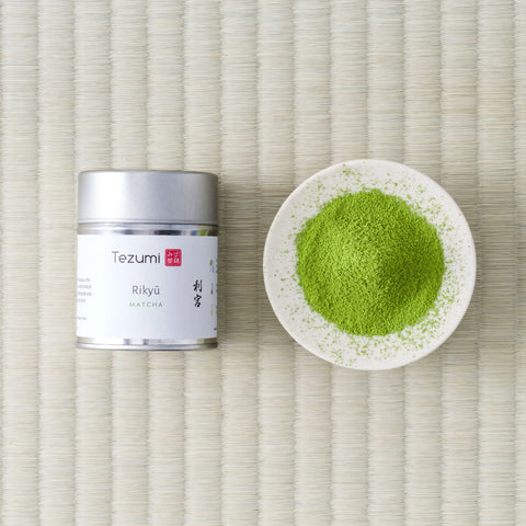 Silver Tezumi tin of Rikyu matcha beside a small dish of green matcha powder on a tatami mat.