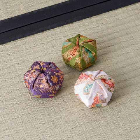 Three Tezumi silk kogo incense containers in green, purple and blush brocade grouped on a tatami mat beside a dark cloth border