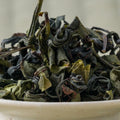 Large green-brown twisted Takachiho oolong leaves piled in a ceramic bowl, side view.
