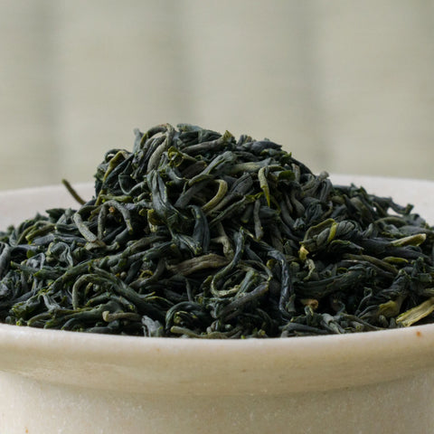 Dark green curled Takachiho Tokujo kamairicha leaves piled in a ceramic bowl, side view.