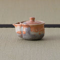 Reddish shino-glazed handleless teapot (houhin) with lid and spout, side view on a tatami mat.