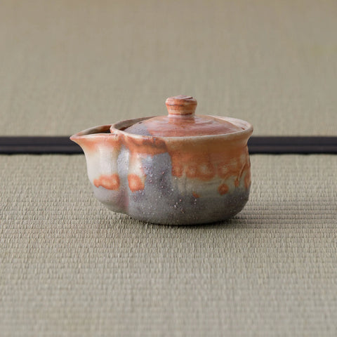 Reddish shino-glazed handleless teapot (houhin) with lid and spout, side view on a tatami mat.