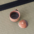Reddish shino-glazed houhin with its lid set beside it, revealing the dark interior, on a tatami mat.