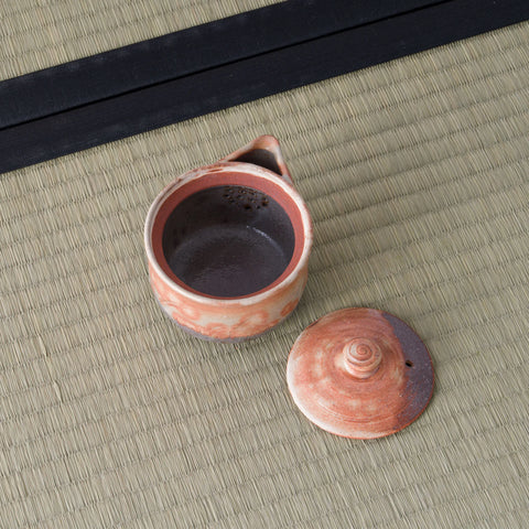 Reddish shino-glazed houhin with its lid set beside it, revealing the dark interior, on a tatami mat.