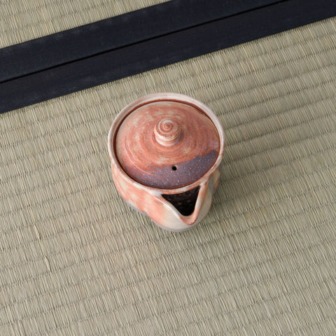 Overhead view of a reddish shino-glazed houhin showing its lid and pouring spout, on a tatami mat.