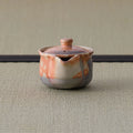 Reddish shino-glazed handleless teapot (houhin) with lid, side view showing the spout, on a tatami mat.