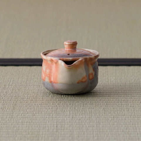 Reddish shino-glazed handleless teapot (houhin) with lid, side view showing the spout, on a tatami mat.